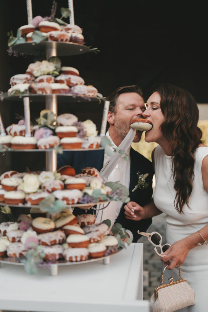 Bruidspaar geniet samen van een feestelijke donut-taart tijdens hun bruiloft. Spontane en speelse trouwfotografie door TwentyTwelve Photography