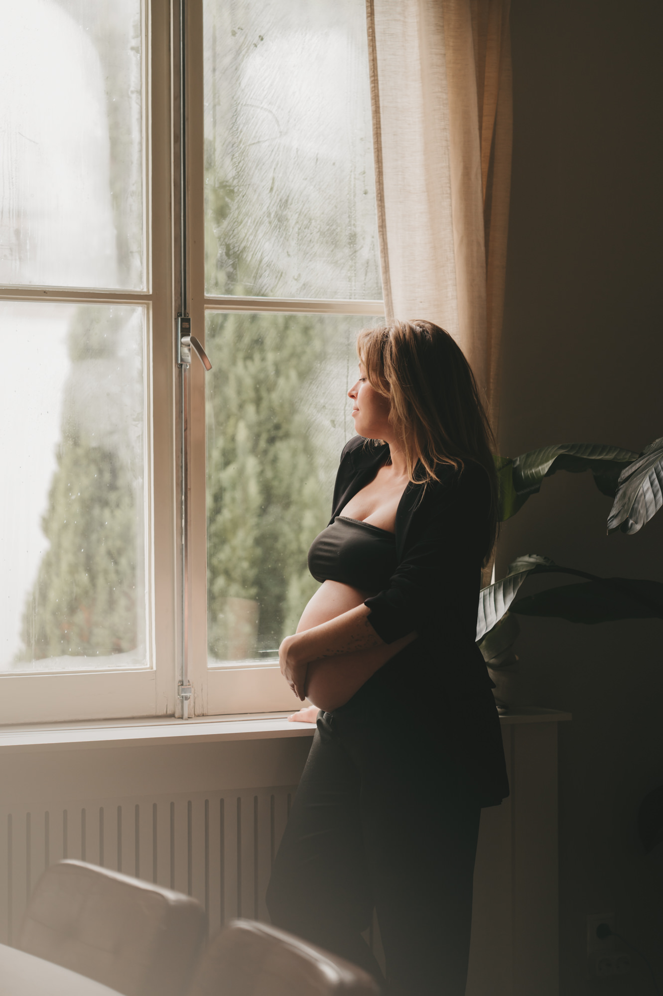 Artistieke zwangerschapsfotografie - zwangere vrouw in eigen woonkamer voor het raam